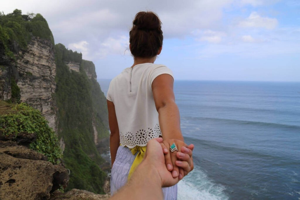 Vizual momka koji drži devojku za ruku na litici sa koje se pruža pogled na more